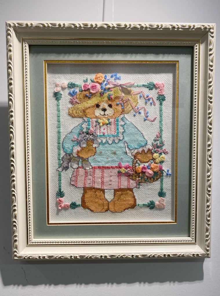 a cross stitch of a teddy bear in a  dress. the teddy bear is holding a basket of flowers and a grey cat.