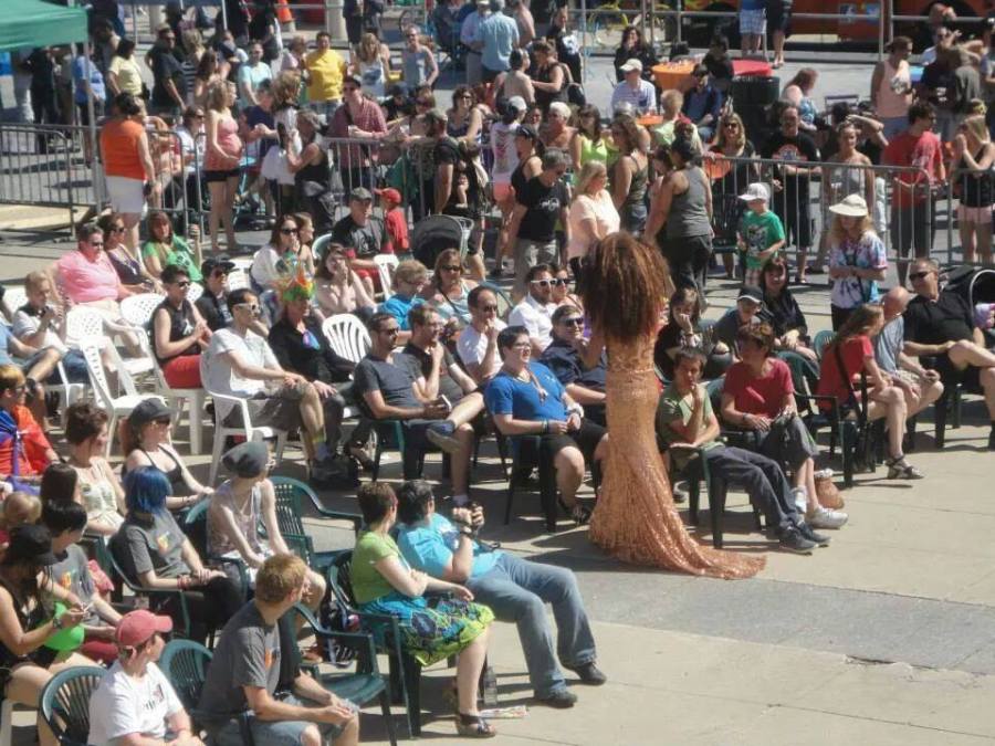 A photo taken in front of city hall where a large group of people of all ages watches a performance by Miss Drew who wears a peach coloured gown and a gigantic curly wig.