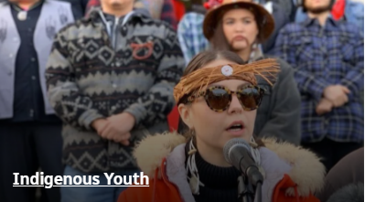 Indigenous youth with individual in sunglasses speaking into a microphone at what appears to be a gathering of people